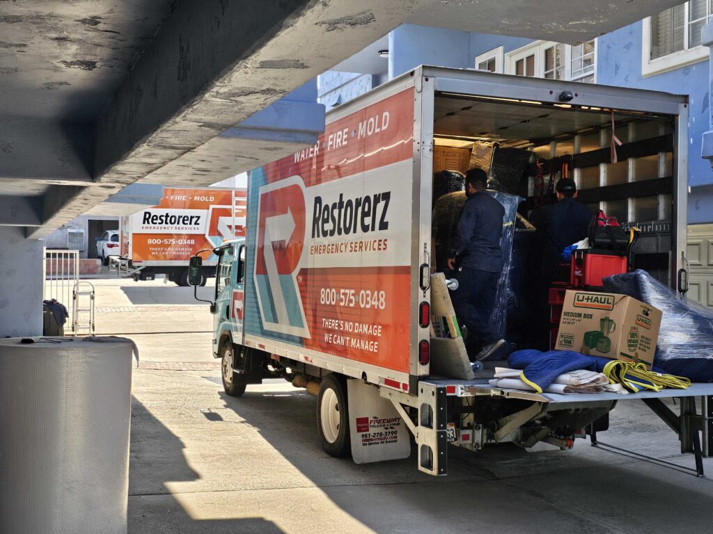 Workers unload equipment from a Restorez emergency services truck parked outside a building, with another Restorez truck visible in the background.