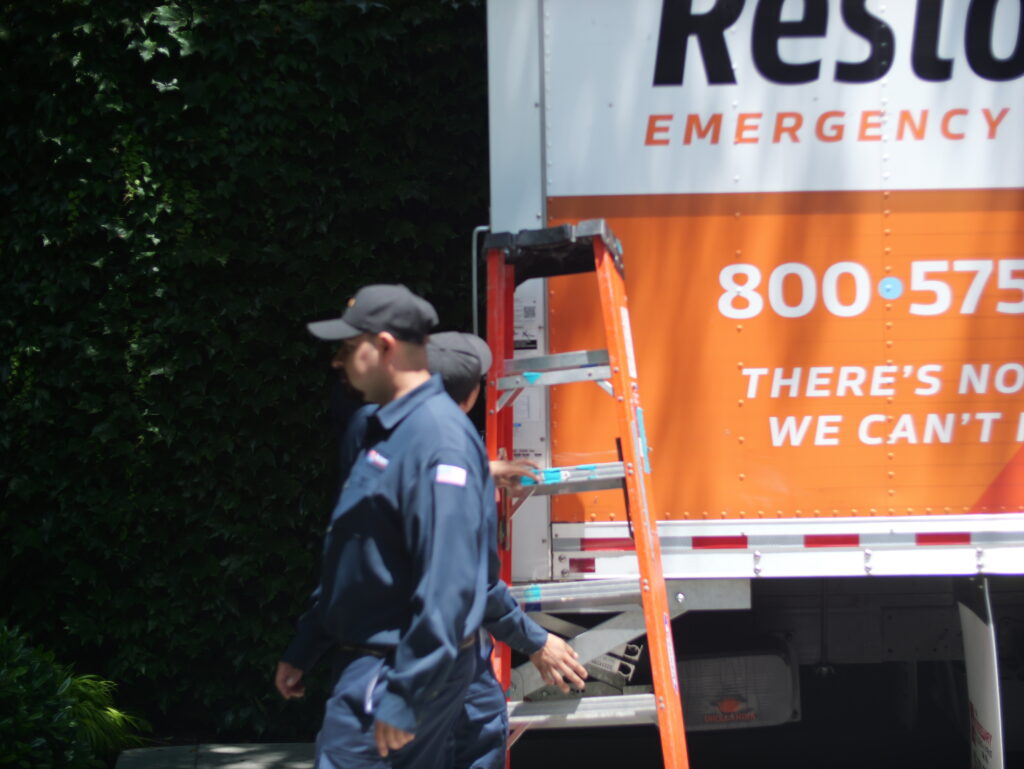Two Restorerz workers in dark uniforms walk past an emergency response truck with a ladder leaning against it.