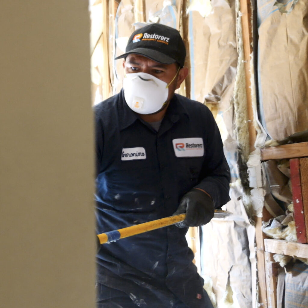 A Restorerz worker wearing a mask, gloves, and a uniform cleans or renovates a damaged building with exposed insulation and debris.
