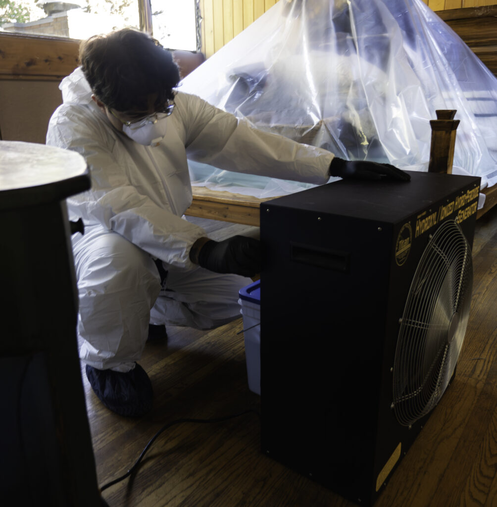A person in full protective gear operates a large dehumidifier or air purifier near covered furniture inside a room with wooden floors.