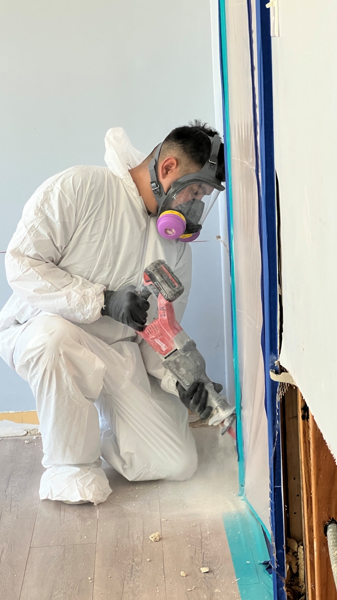 A person wearing protective gear and a respirator uses a power saw to cut into the wall near a floor, creating dust.