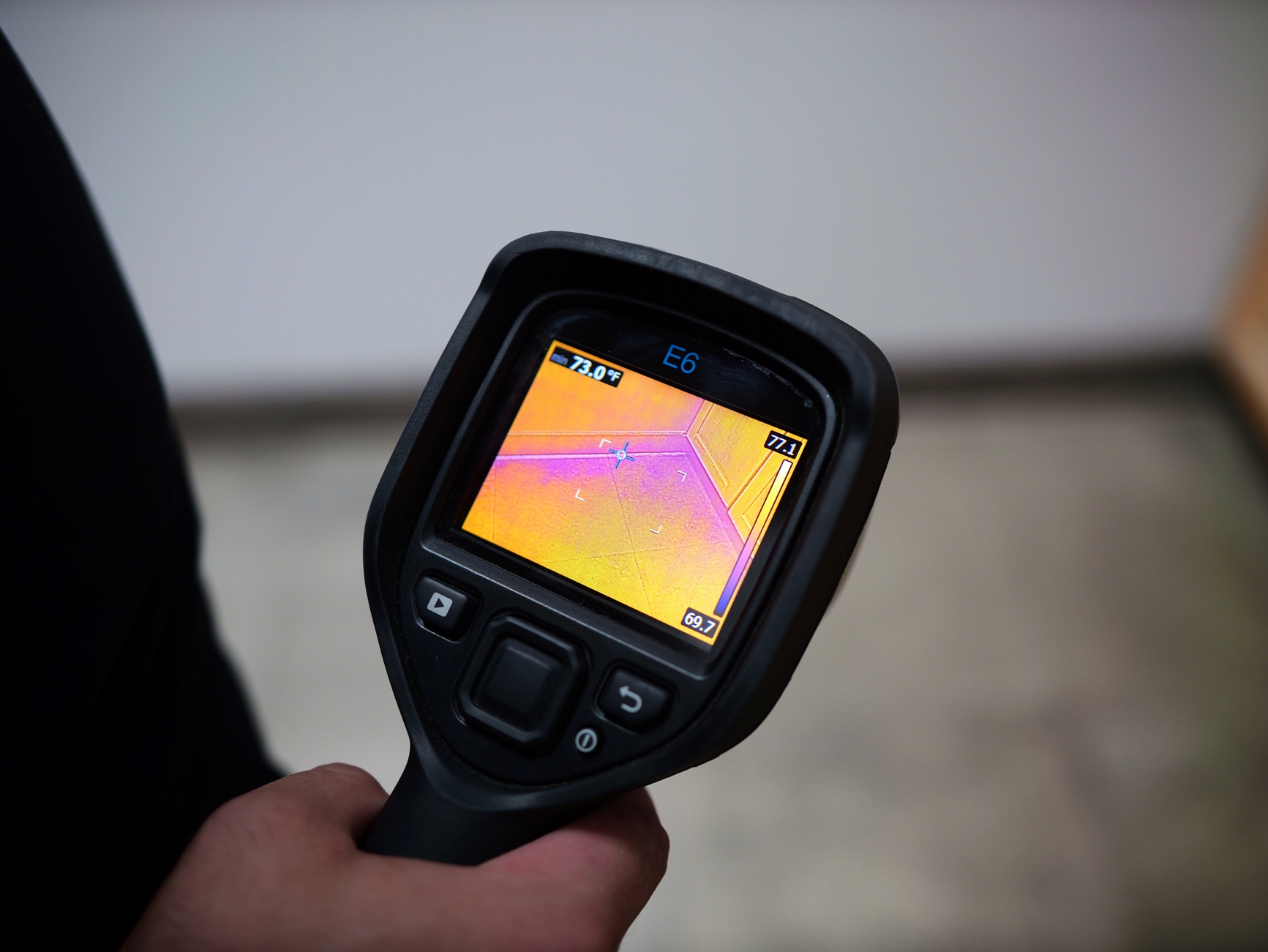 A person holds a thermal imaging camera displaying a heat map of a room&rsquo;s corner, with temperature readings shown on the screen.
