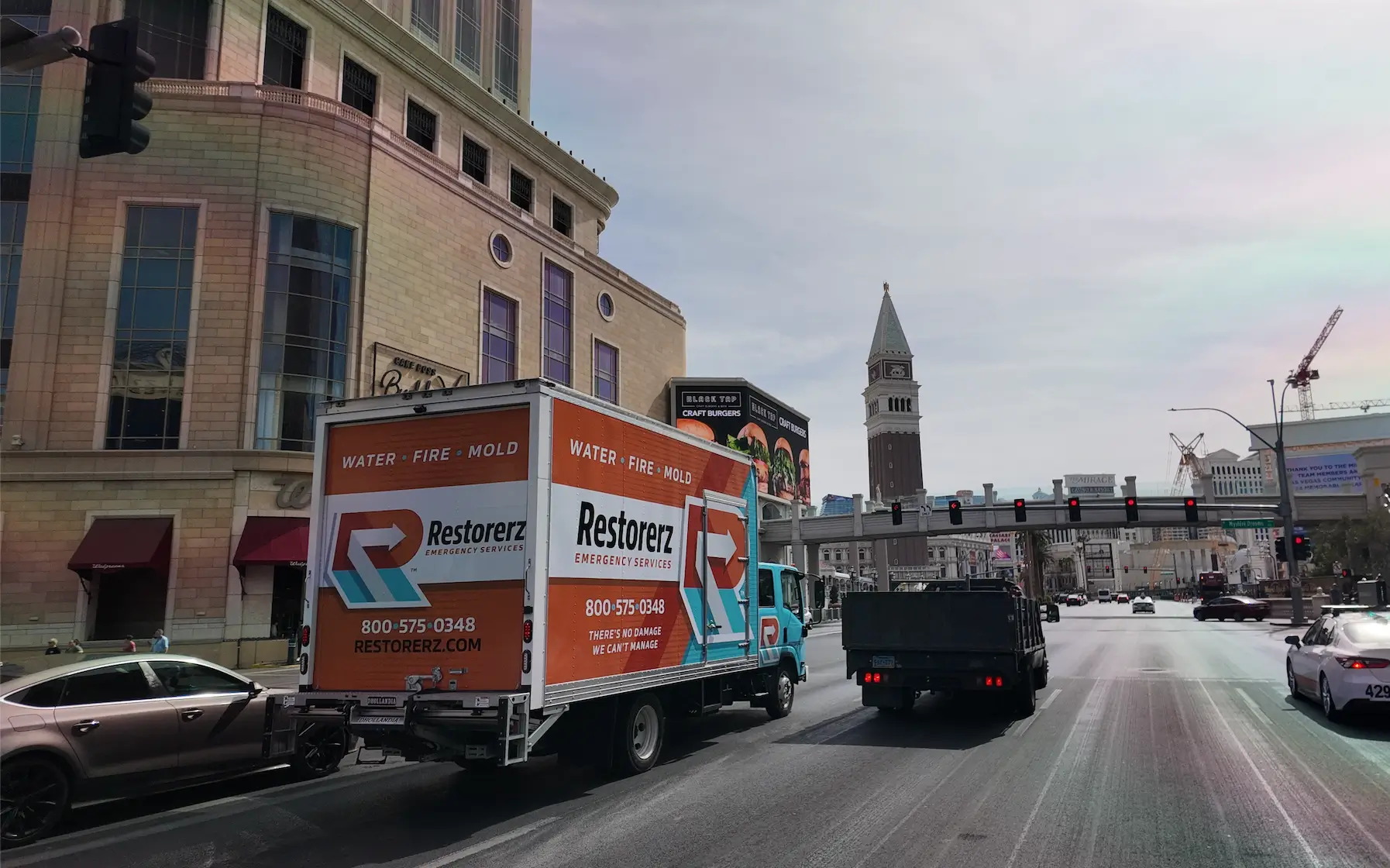 A Restorerz emergency services truck drives on a city street, passing buildings and a clock tower under a partly cloudy sky.
