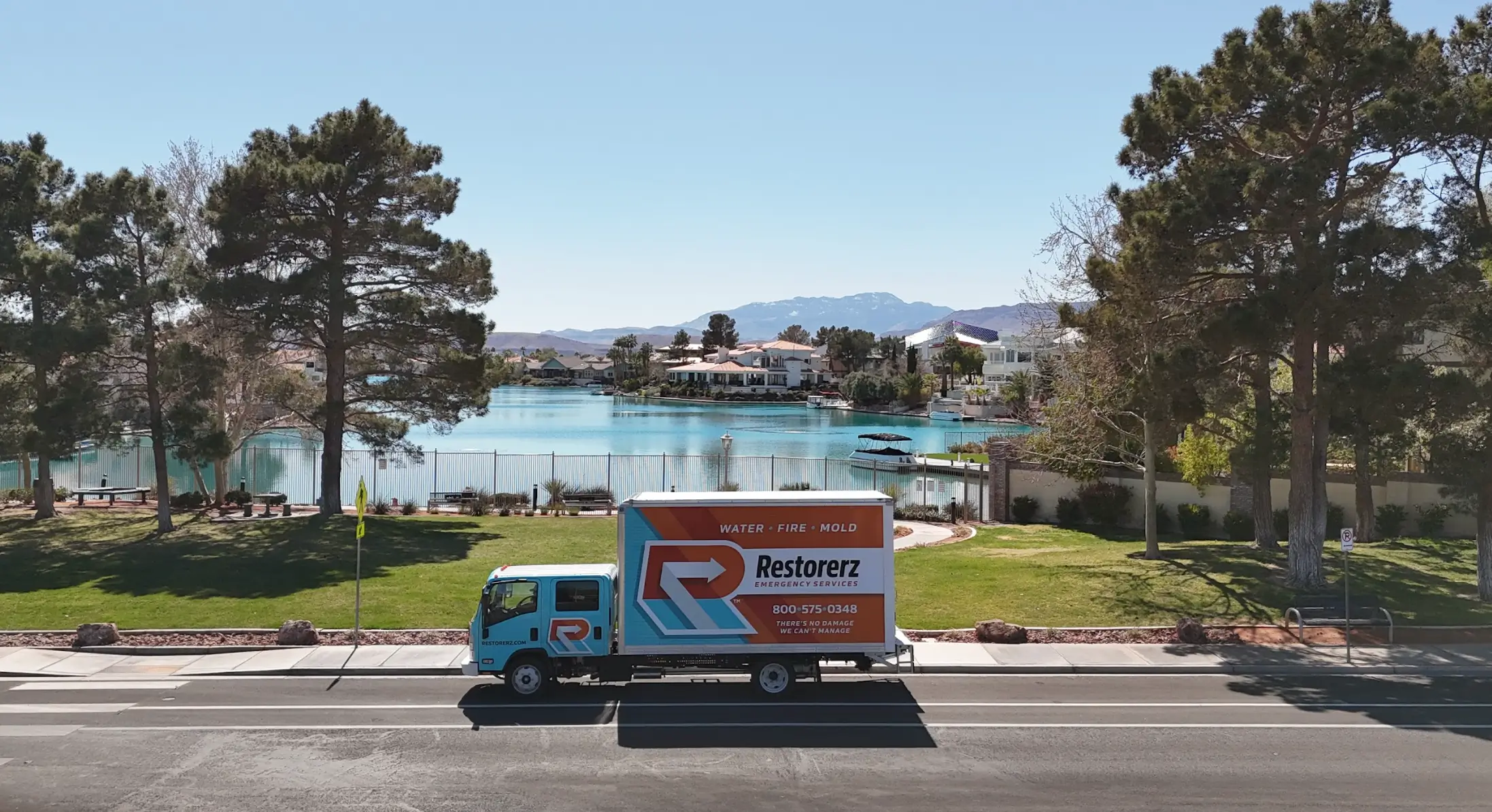 A Restorez service truck is parked on a road in front of a lake, with trees, grass, houses, and mountains visible in the background on a clear day.