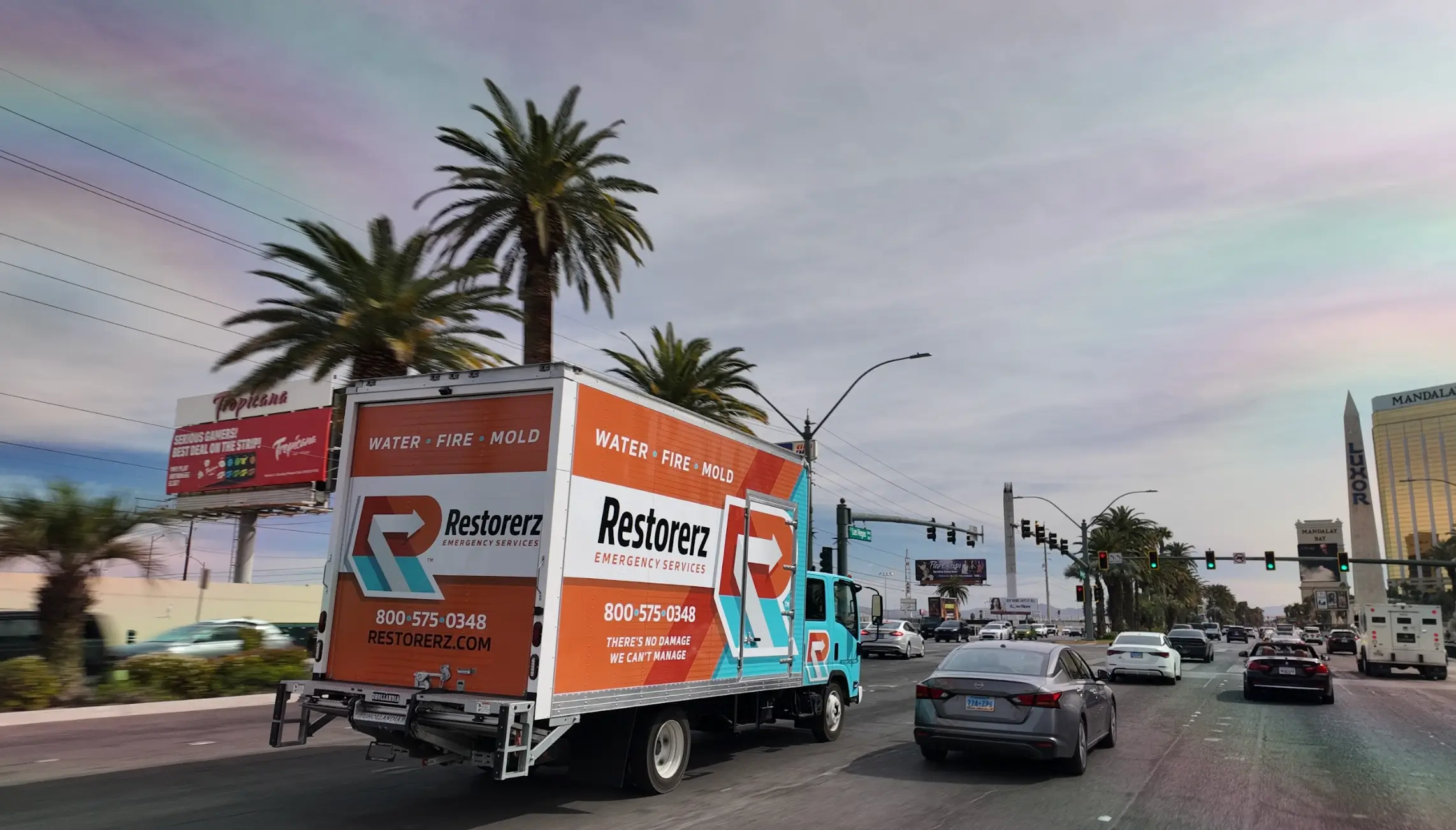 A Restorerz emergency restoration truck drives down a city street lined with palm trees and traffic under a cloudy sky.