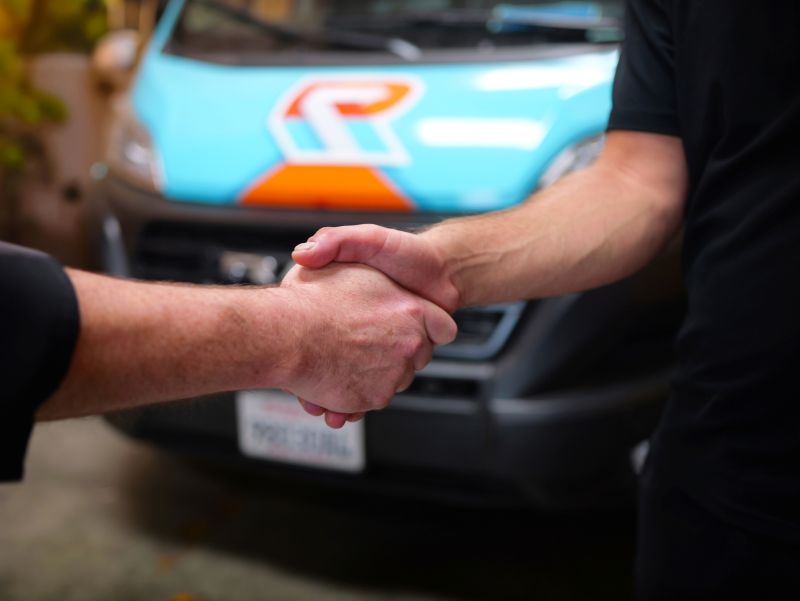 Two people shake hands in front of a blue and orange vehicle, partially visible in the background.