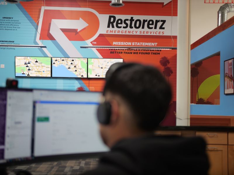 A person with headphones sits at a computer in an office with a "Restorerz Emergency Services" sign and digital maps on the wall.